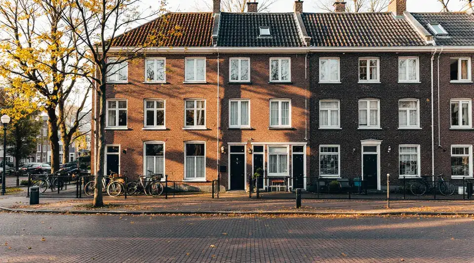 Een rij huizen van rode baksteen in de ochtendzon, met fietsen tegen het hek en herfstbladeren op de klinkers